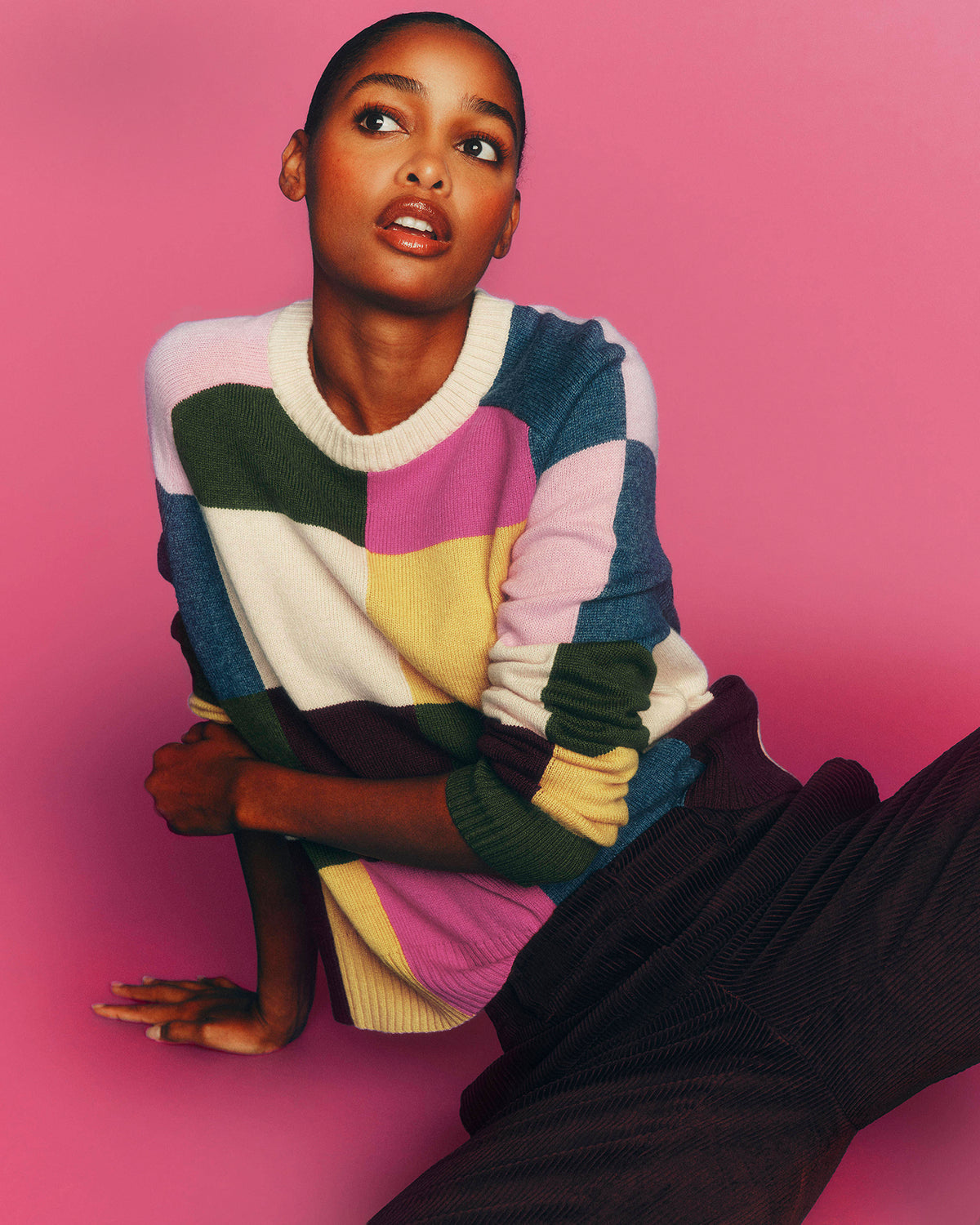 A model wearing a colorful patchwork knit sweater and dark corduroy pants, posed against a solid pink background.
