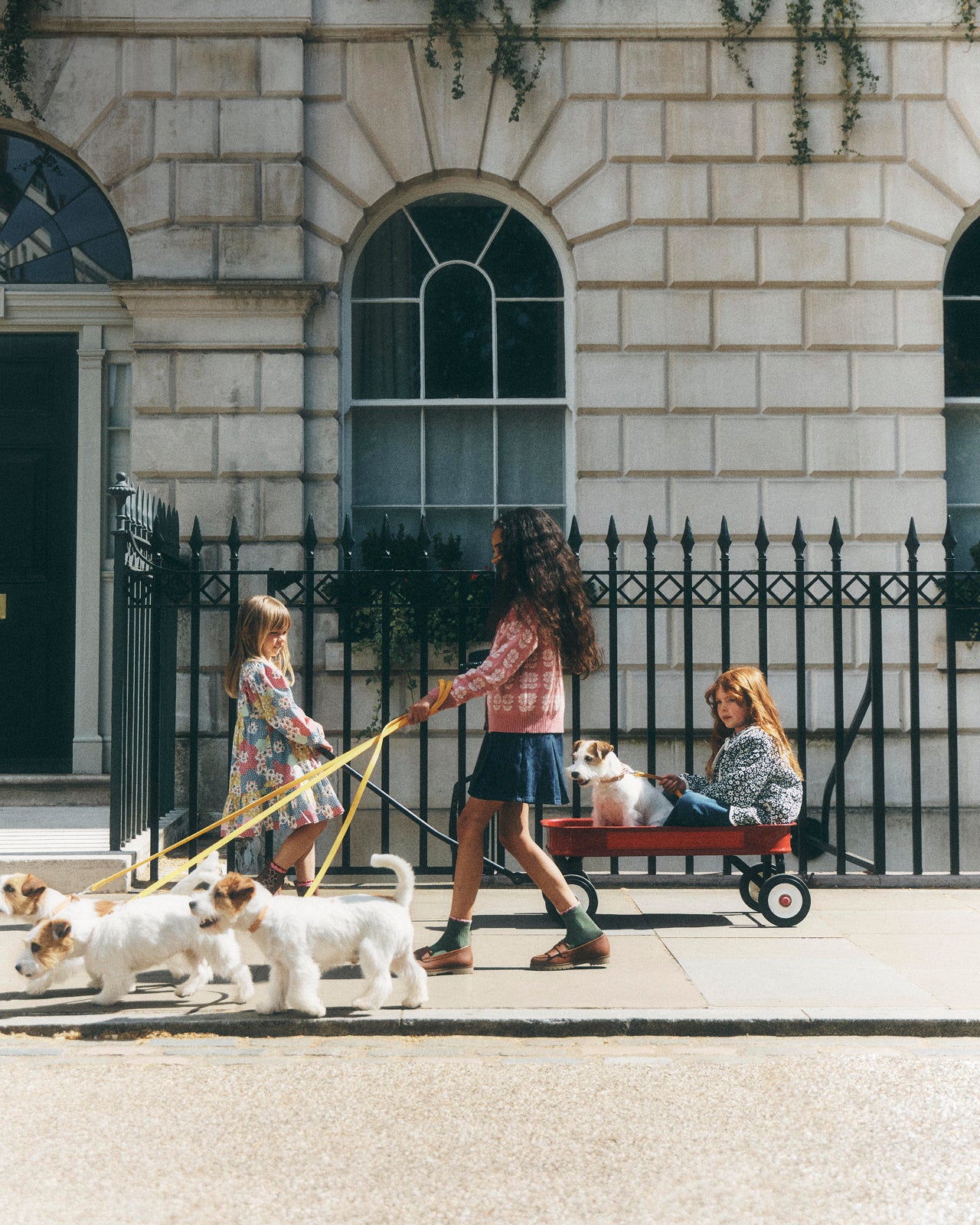 A woman walks two small dogs on leashes while sitting on a red wagon with a child, set against a classic stone building and black iron fence.