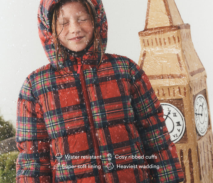 A child wearing a red and black plaid hooded puffer jacket stands outdoors in the rain next to a large wooden clock tower prop. White text overlays at the bottom highlight product features like water resistance, cozy ribbed cuffs, soft lining, and heavy wadding.