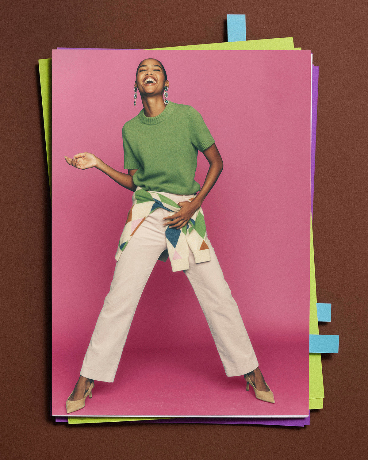A fashion editorial-style photo print featuring a woman in a green short-sleeve top and white pants posing against a bright pink backdrop, surrounded by stacked colorful paper sheets on a brown background.