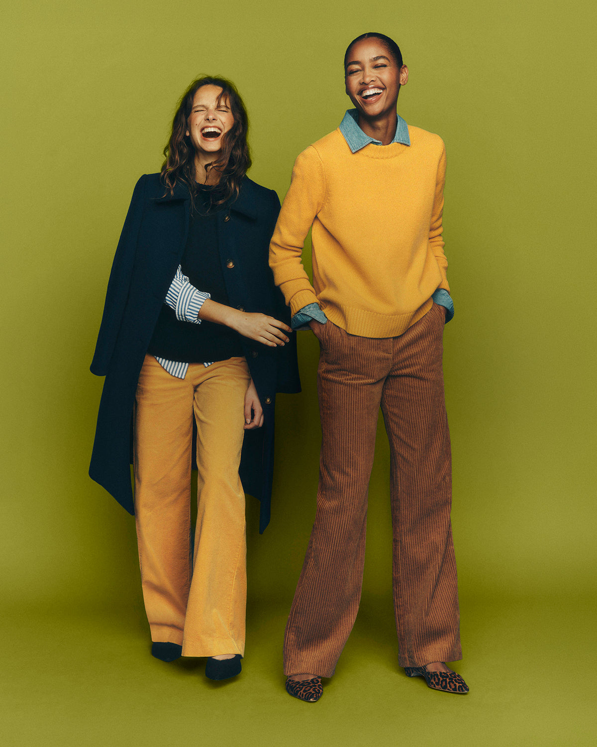 A man and a woman modeling coordinated outfits against a solid olive green studio background. The woman wears a dark navy cape over a black top and mustard yellow flared pants.