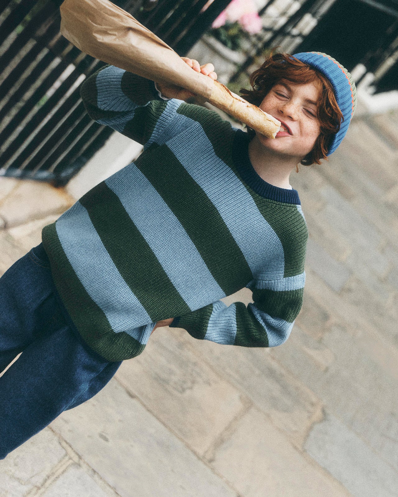 A child wearing a green and light blue striped knit sweater, dark blue pants, and a blue beanie, holding a brown paper bag near their face while standing outdoors on stone steps.