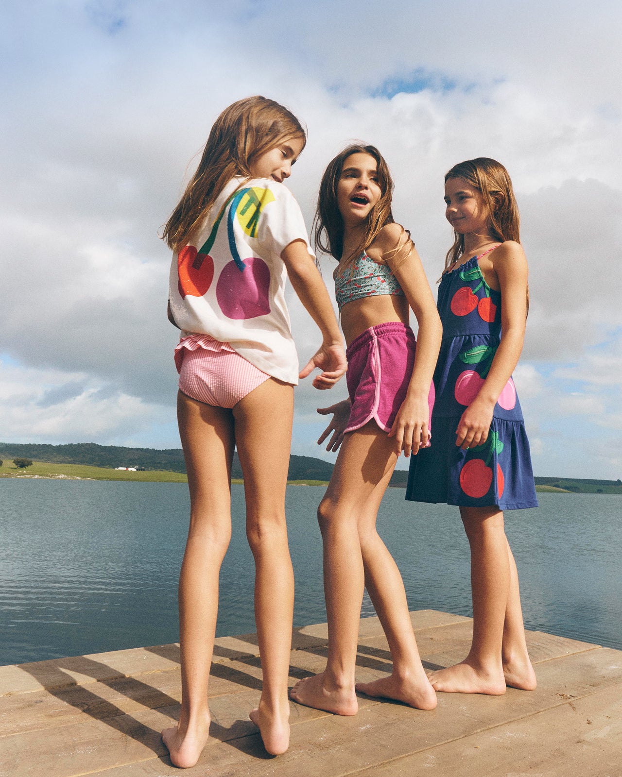 Three young girls standing on a dock by a lake, wearing cherry-themed clothing.