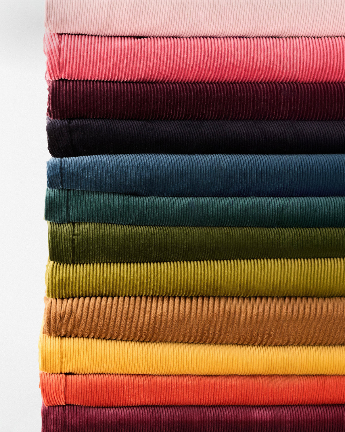 A vertical stack of folded corduroy fabric swatches in multiple colors, including pink, burgundy, navy, teal, green, olive, mustard, tan, yellow, orange, and red, shown against a plain white background.