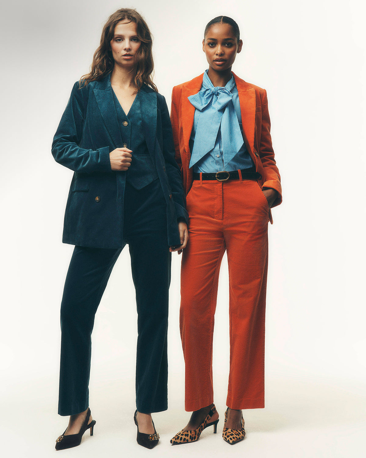 Two models posing in matching tailored suits. The model on the left wears a dark teal velvet blazer and trousers set with black heels.