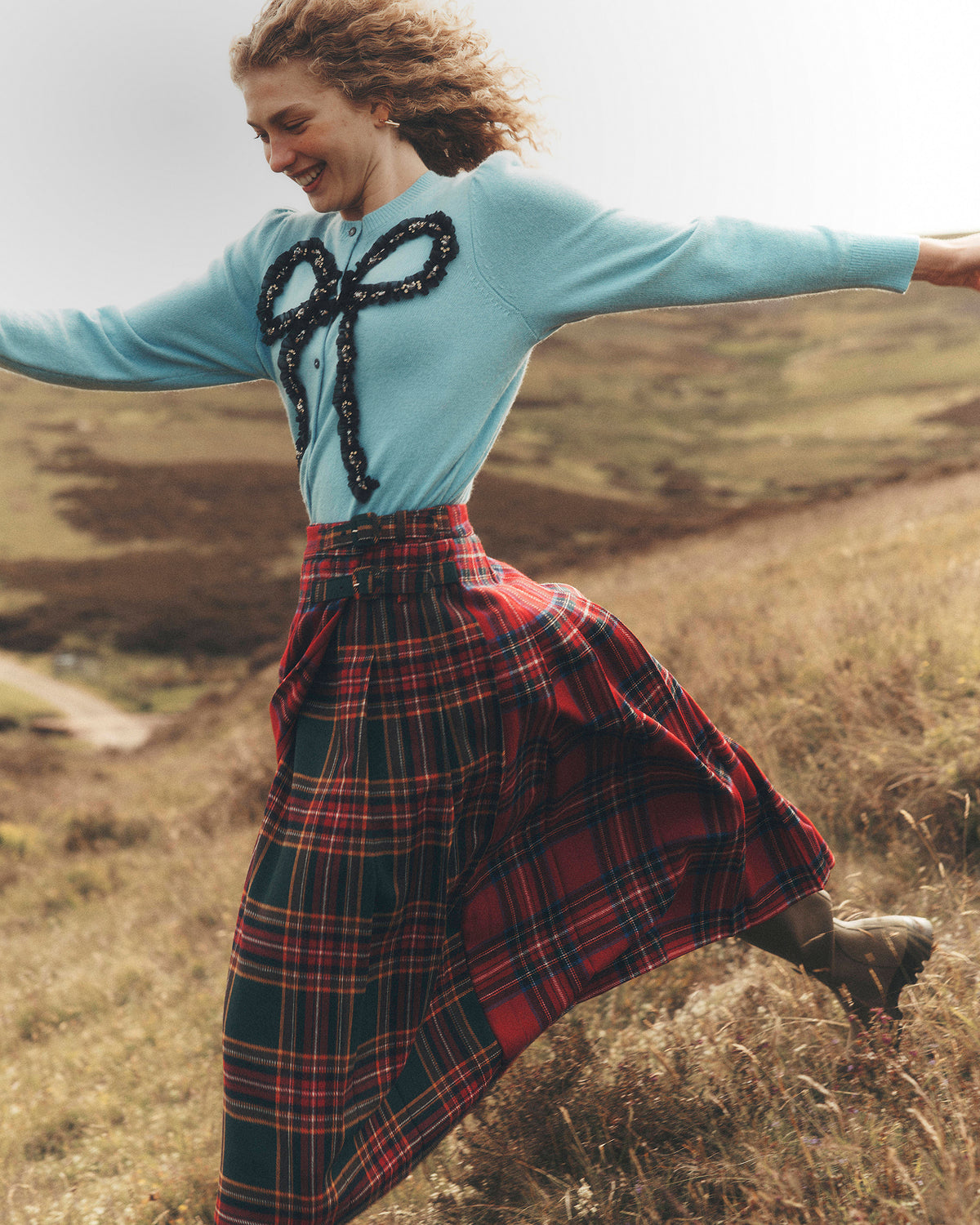 A woman outdoors in a grassy field wearing a light blue knit sweater with a black patterned bow detail on the chest and a red tartan plaid skirt.