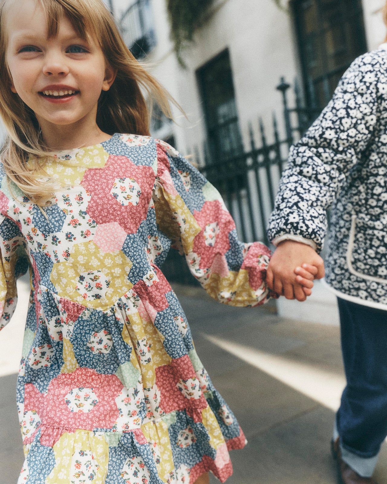 A young girl smiling and holding hands with another child outdoors. The girl wears a long-sleeve patchwork dress with a floral and geometric print in pink, yellow, blue, and white.