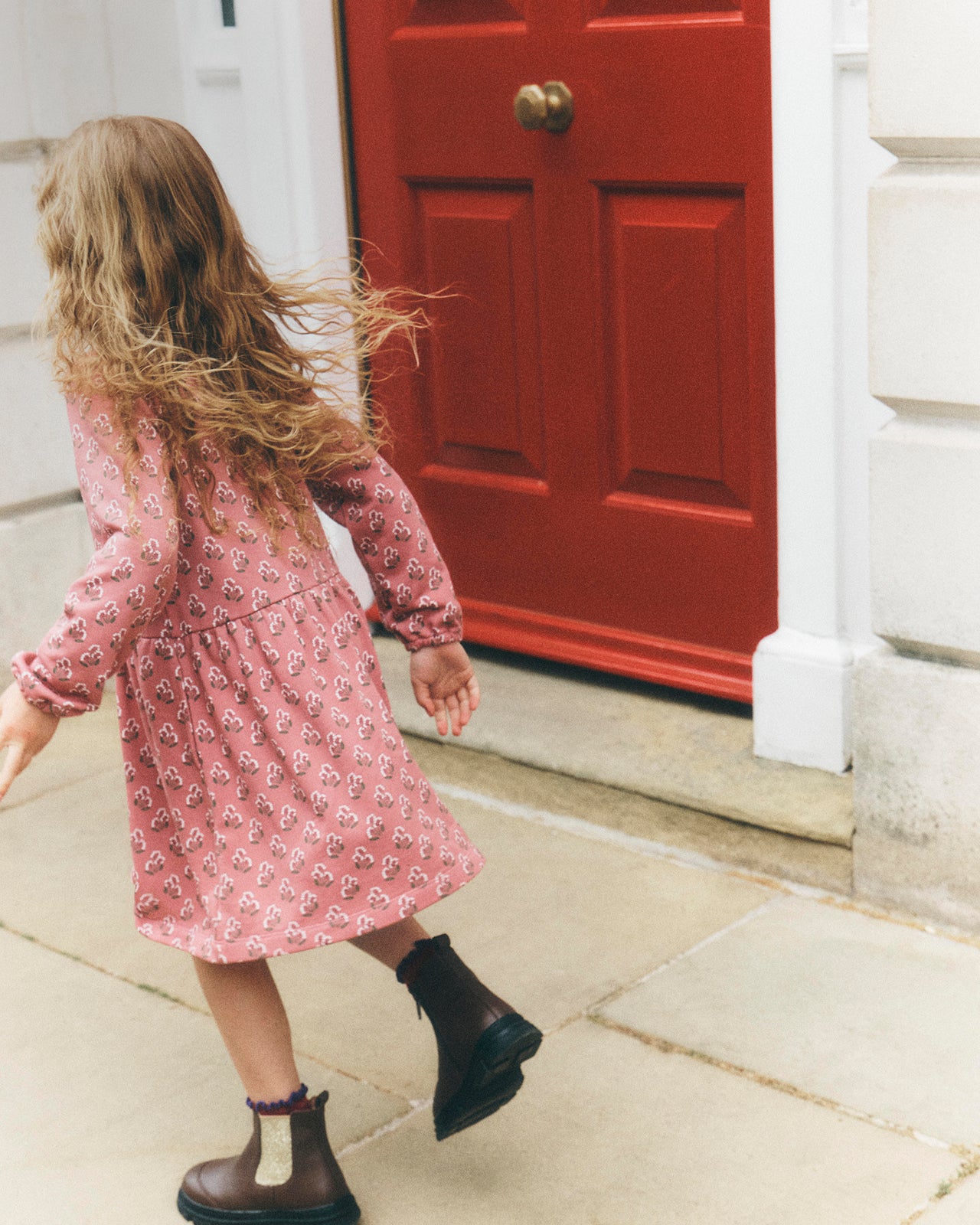 A young girl with long blonde hair is seen from behind, wearing a pink floral long-sleeve dress and dark brown ankle boots, walking on a stone sidewalk towards a red door.