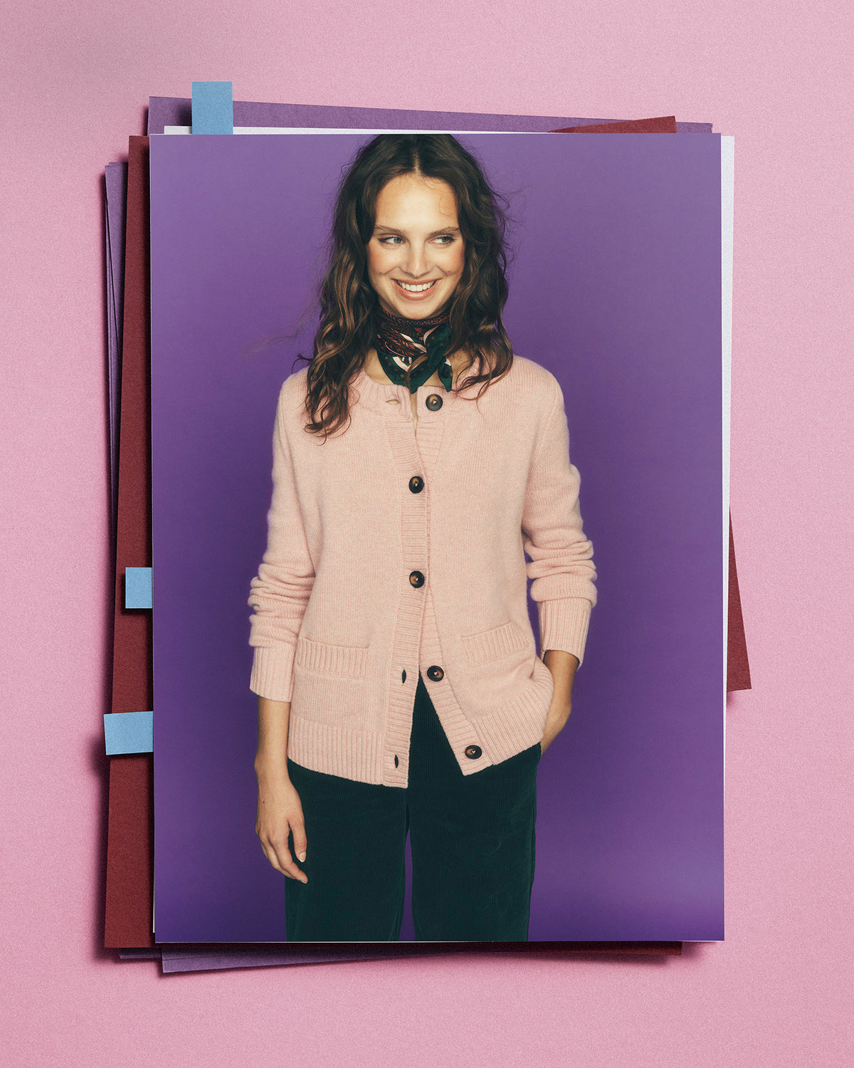 A printed photo or poster showing a smiling woman wearing a light pink button-front cardigan and dark pants, set against a purple background and photographed on a pink surface with stacked paper sheets around it.