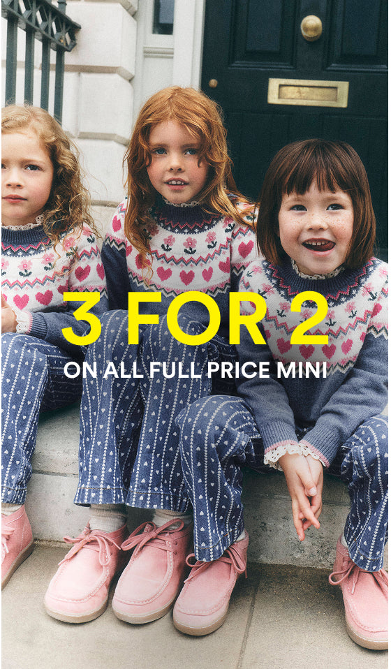 Three young girls sitting on steps in front of a dark door, wearing matching knit sweaters and trousers in a pink, white, and blue pattern. Large yellow text across the center reads '3 FOR 2' with smaller white text below reading 'ON ALL FULL PRICE MINI'.