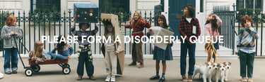 A group of children and two small dogs standing outdoors in front of a building with a black iron fence. The children are holding cardboard boxes and wearing casual clothing, and large white text across the center reads, 'PLEASE FORM A DISORDERLY QUEUE'.