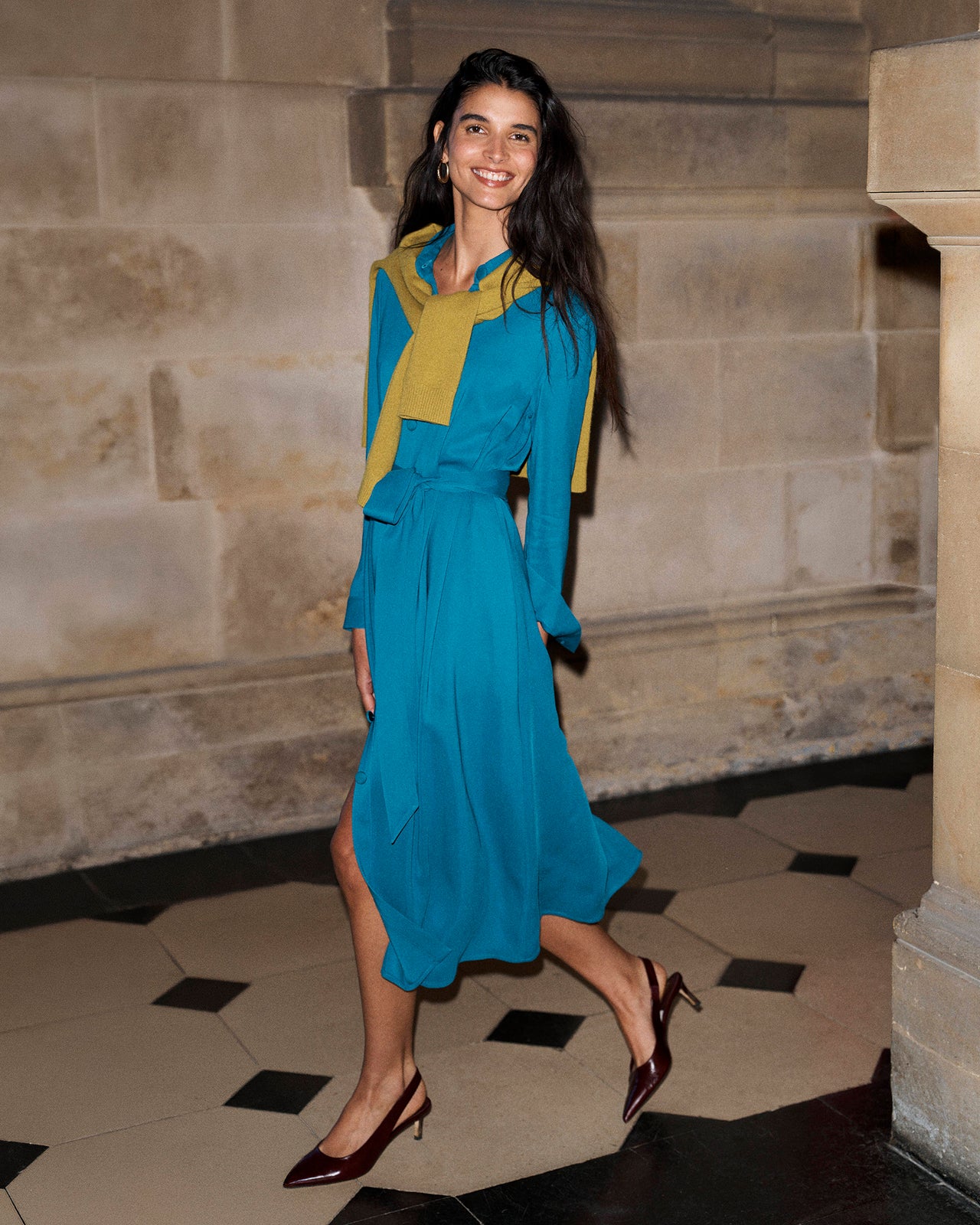 A woman wearing a vibrant teal blue midi dress with a yellow color-blocked collar and cuffs, styled with dark brown pointed-toe heels, posing in front of a stone wall.