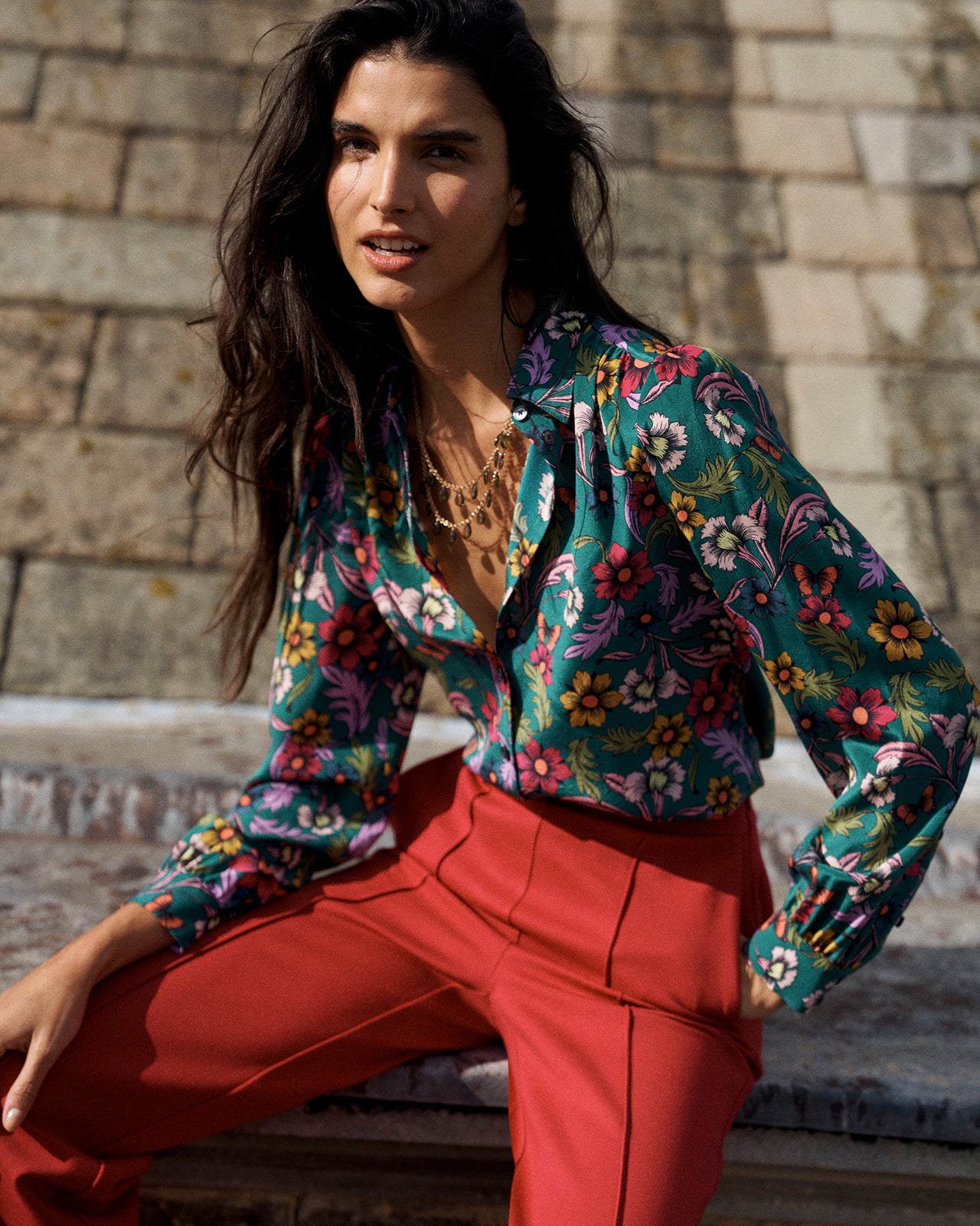 A woman seated outdoors against a stone wall, wearing a long-sleeve floral button-up shirt in teal with multicolored flowers and bright red pants.