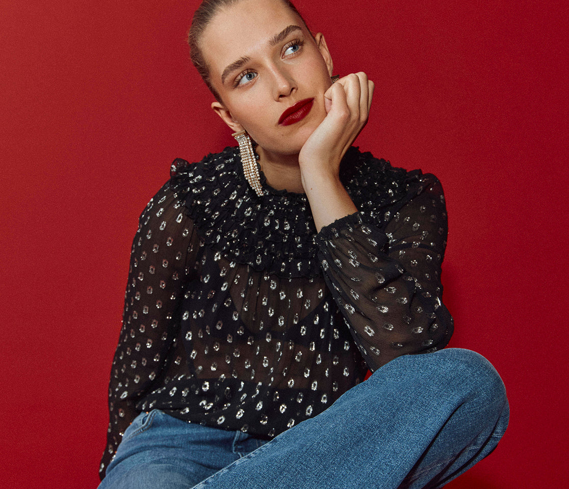 Woman wearing a black patterned blouse and blue jeans against a red background