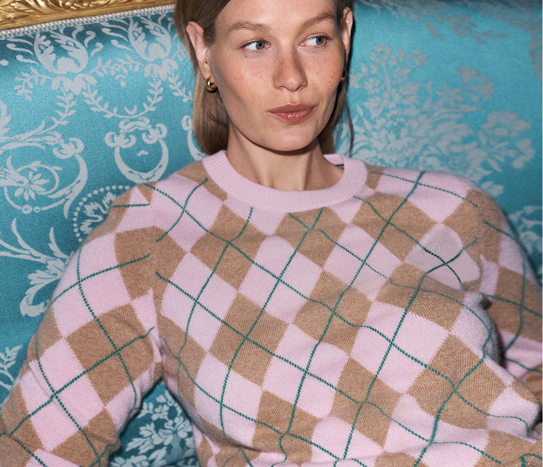 A woman wearing a pink and tan argyle sweater with a thin green grid pattern, seated on a turquoise patterned sofa.