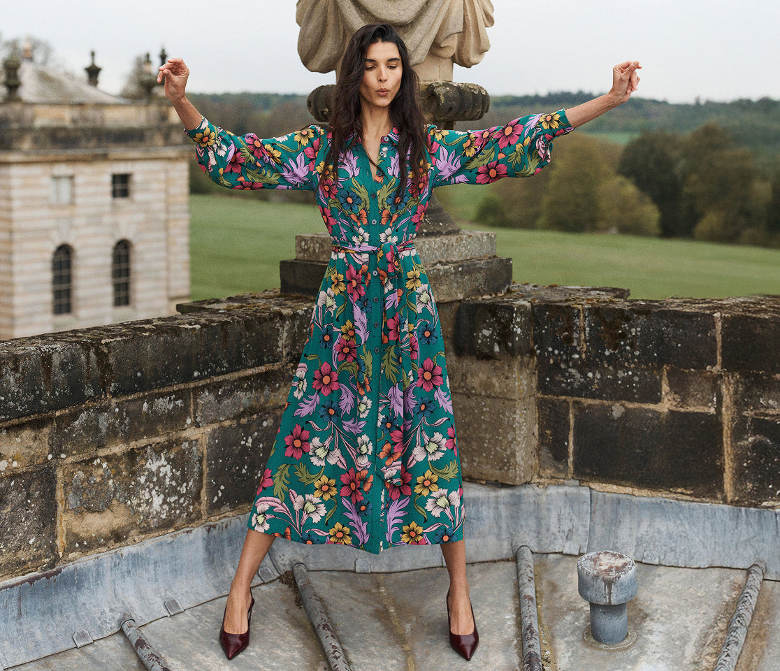 A woman stands on a stone terrace wearing a long-sleeved, knee-length wrap dress with a vibrant floral print on a teal background. The dress features pink, purple, yellow, and white flowers.