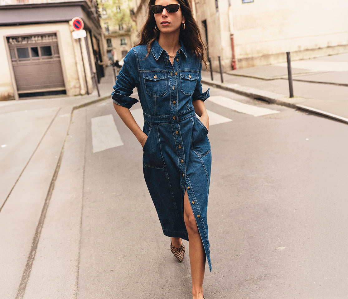 A woman wearing a dark blue denim shirt dress walks down a city street. The dress has a button-front closure, chest pockets, and a high slit on one side.