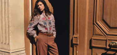 A woman stands in a doorway wearing a light pink floral button-up shirt and brown wide-leg trousers, posed against a warm-toned wooden door and stone wall.