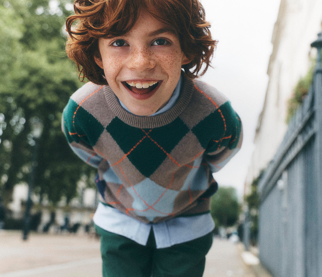 A young boy with curly red hair and freckles smiles broadly while leaning forward on a city street. He is wearing a green, brown, and gray argyle sweater over a light blue collared shirt and dark green pants.