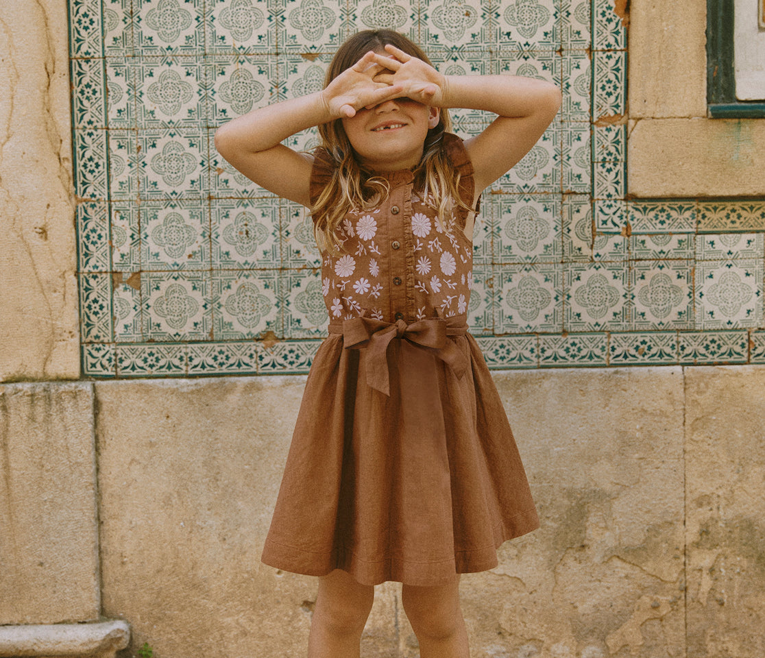 Young girl in a brown dress standing in front of a patterned tile wall.