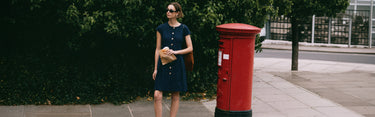 Woman in a blue dress standing next to a red postbox on a sidewalk.