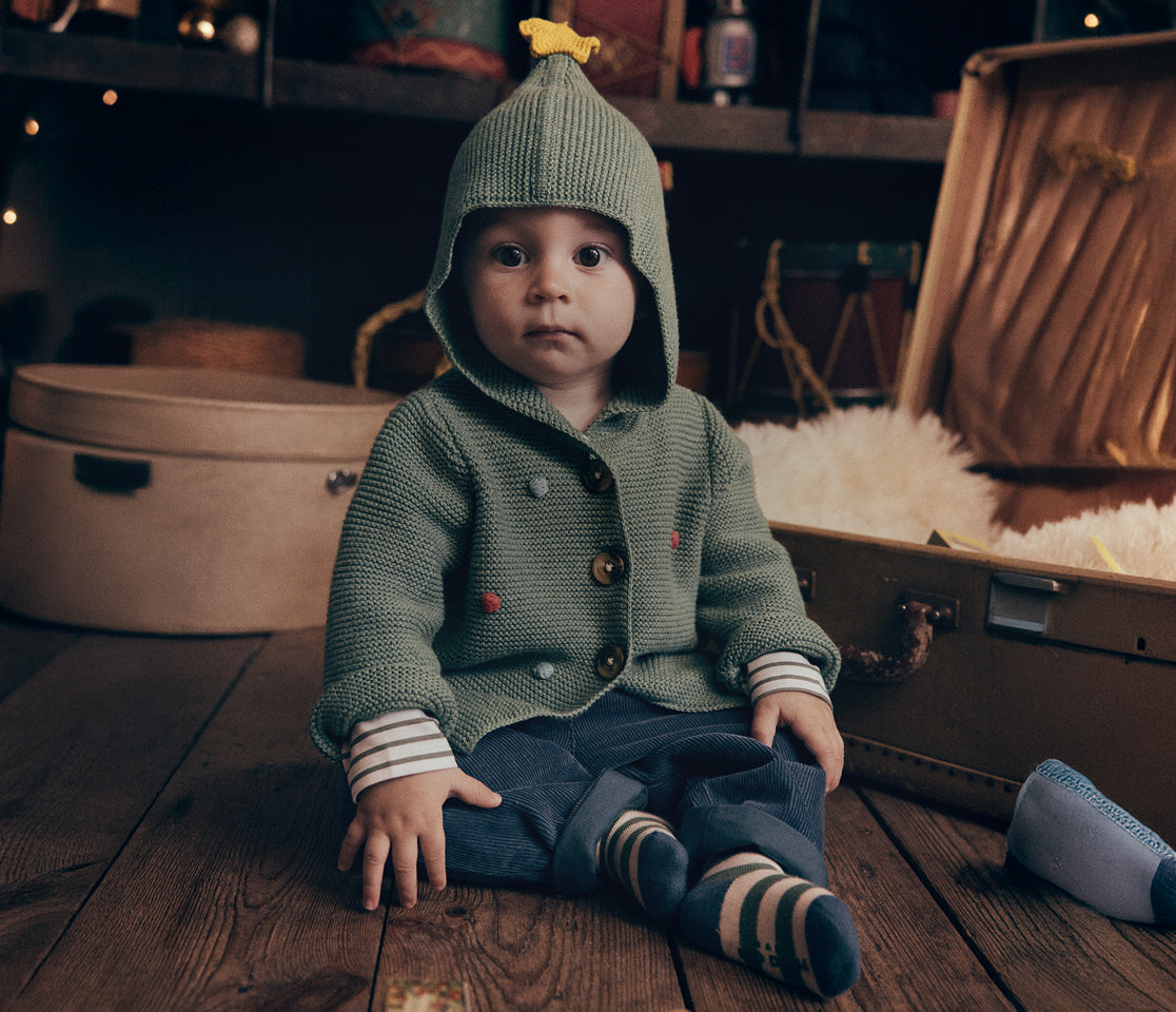 A baby sitting on a wooden floor wearing a green knit hooded cardigan with a yellow bird accent on the hood, dark pants, and striped socks, with vintage suitcases in the background.
