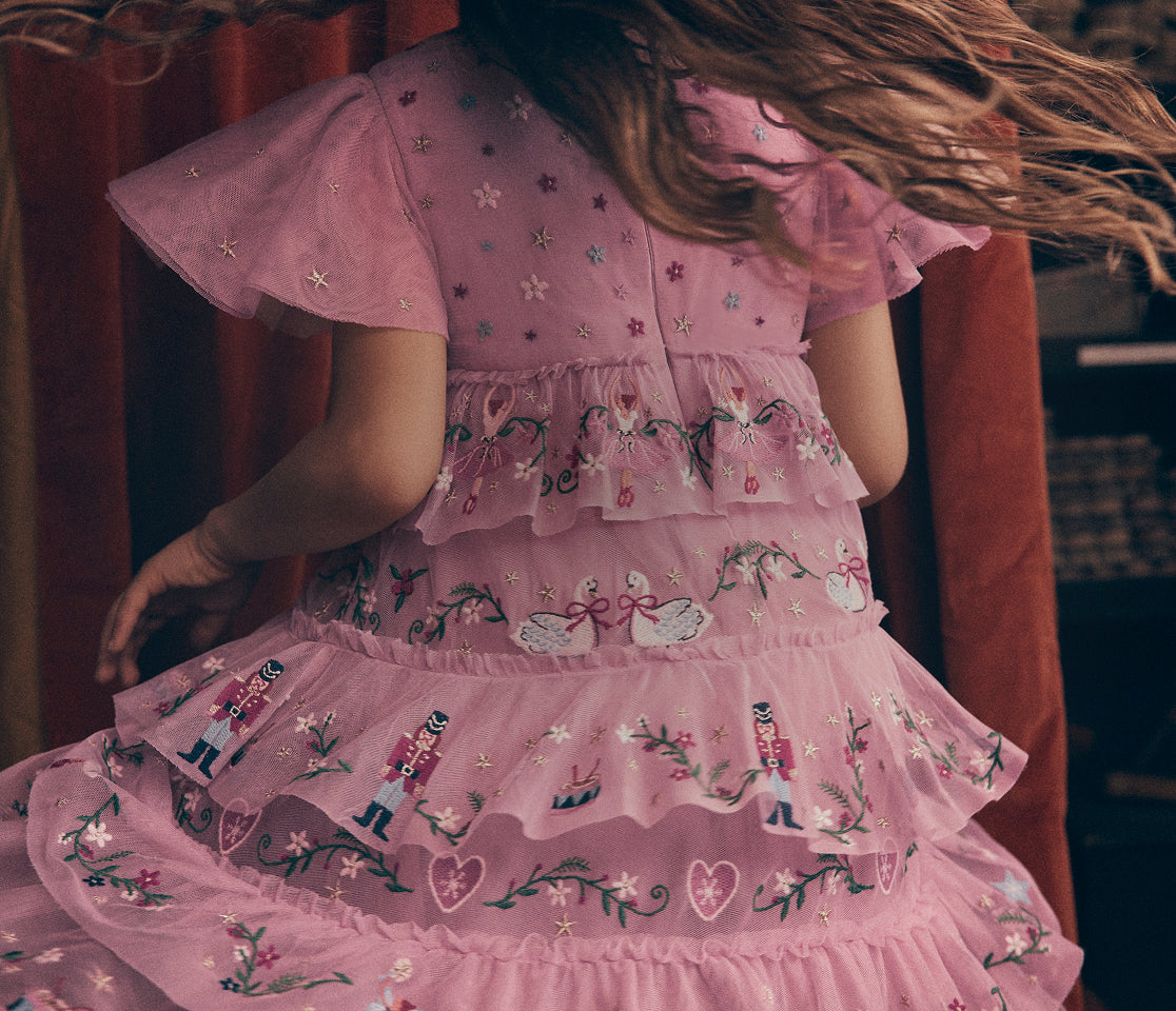 A pink tiered tulle dress with embroidered swans, nutcrackers, and floral motifs, shown from the back on a model with long brown hair.