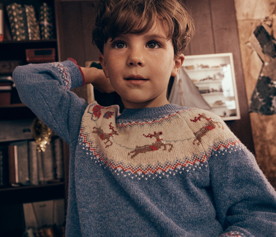 Close-up of a child wearing a blue Fair Isle knit jumper featuring a yoke pattern with reindeer motifs in cream, red and brown. The image is set indoors against a cosy backdrop of bookshelves and rustic wooden details, highlighting the intricate knit design and soft texture of the sweater.