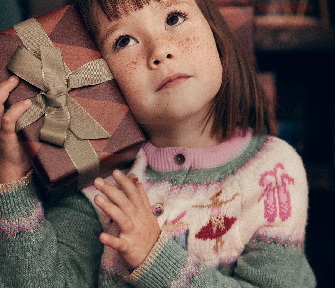 Close-up of a child holding a wrapped gift with a gold ribbon, wearing a Fair Isle knit cardigan in soft pink, cream and sage green with festive motifs including ballet shoes and dancers. The image highlights the detailed knit pattern and cosy texture against a warm indoor background.