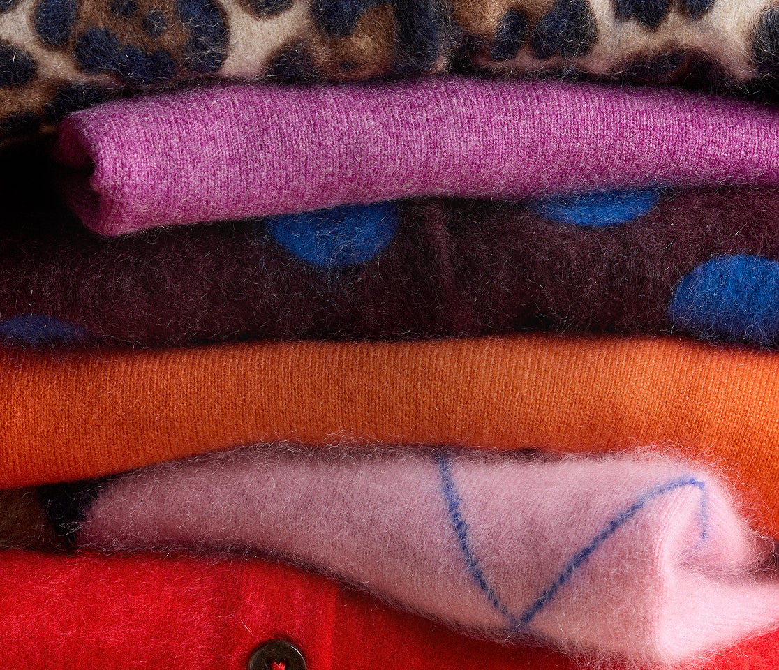 A stack of colourful folded knitwear featuring leopard print, teal, pink, burgundy, orange and red fabrics with soft woolly textures and button details.