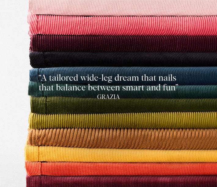 A stack of folded ribbed fabric in multiple colors, including pink, burgundy, navy, teal, olive green, mustard yellow, orange, and deep red, with a white quote overlaid in the center reading, "A tailored wide-leg dream that nails that balance between smart and fun" GRAZIA.