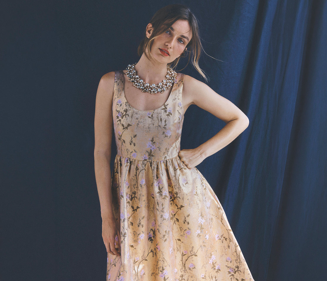 A woman models a sleeveless beige midi dress with a delicate purple and green floral pattern, styled with a chunky silver statement necklace against a dark blue backdrop.