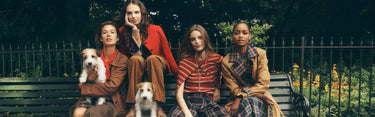 Four models sitting on a park bench with two small dogs, wearing coordinated earth-toned outfits including brown, rust red, and plaid patterns against a dark garden background.