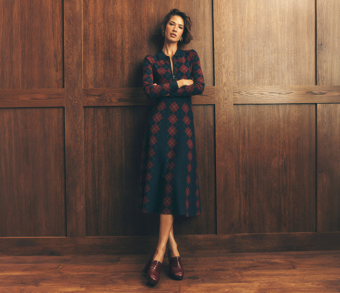 A woman stands against a dark wood-paneled wall wearing a long-sleeve midi dress with a red and black argyle pattern, paired with dark brown leather loafers.