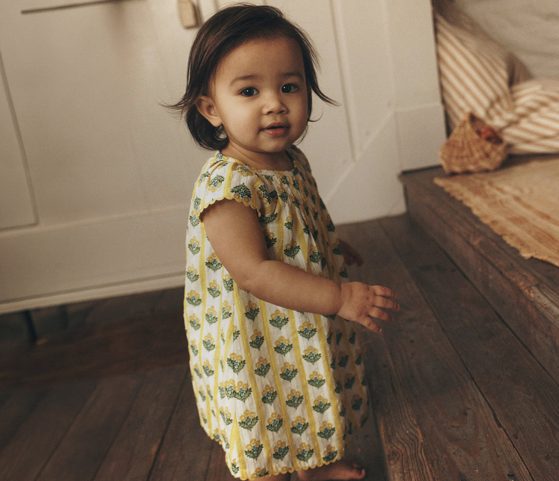 Child in a yellow and green patterned dress standing on a wooden floor.