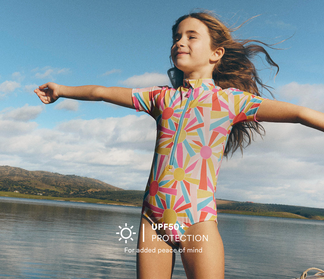 Child in a colorful swimsuit standing by a lake with UPF50+ protection label.