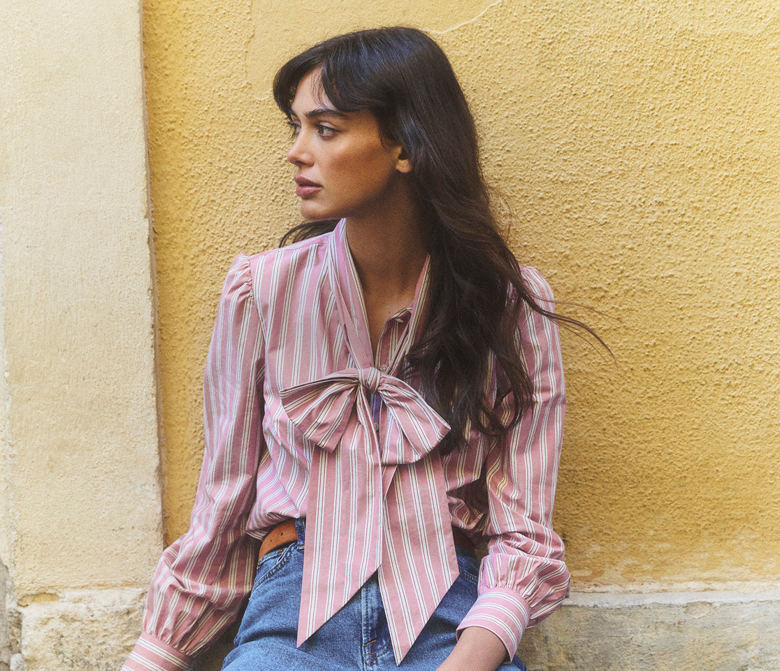 Woman wearing a pink striped shirt with a bow tie against a yellow wall.