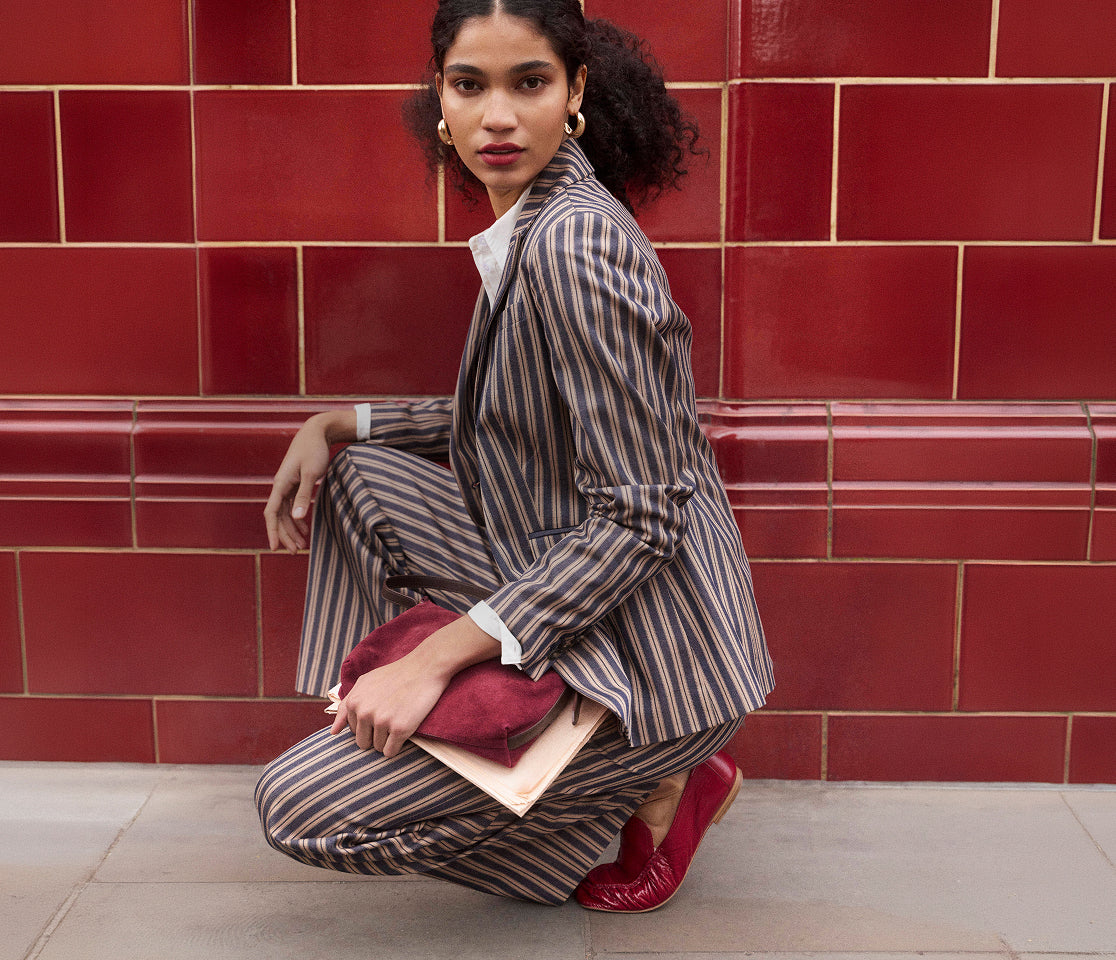 A model crouches in front of a red tiled wall wearing a brown and beige vertically striped blazer and matching trousers, styled with a white collared shirt, a burgundy clutch, and red loafers.