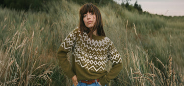 A woman stands in a grassy field wearing a long-sleeve knit sweater with a geometric Fair Isle pattern in olive green and cream, paired with blue jeans.