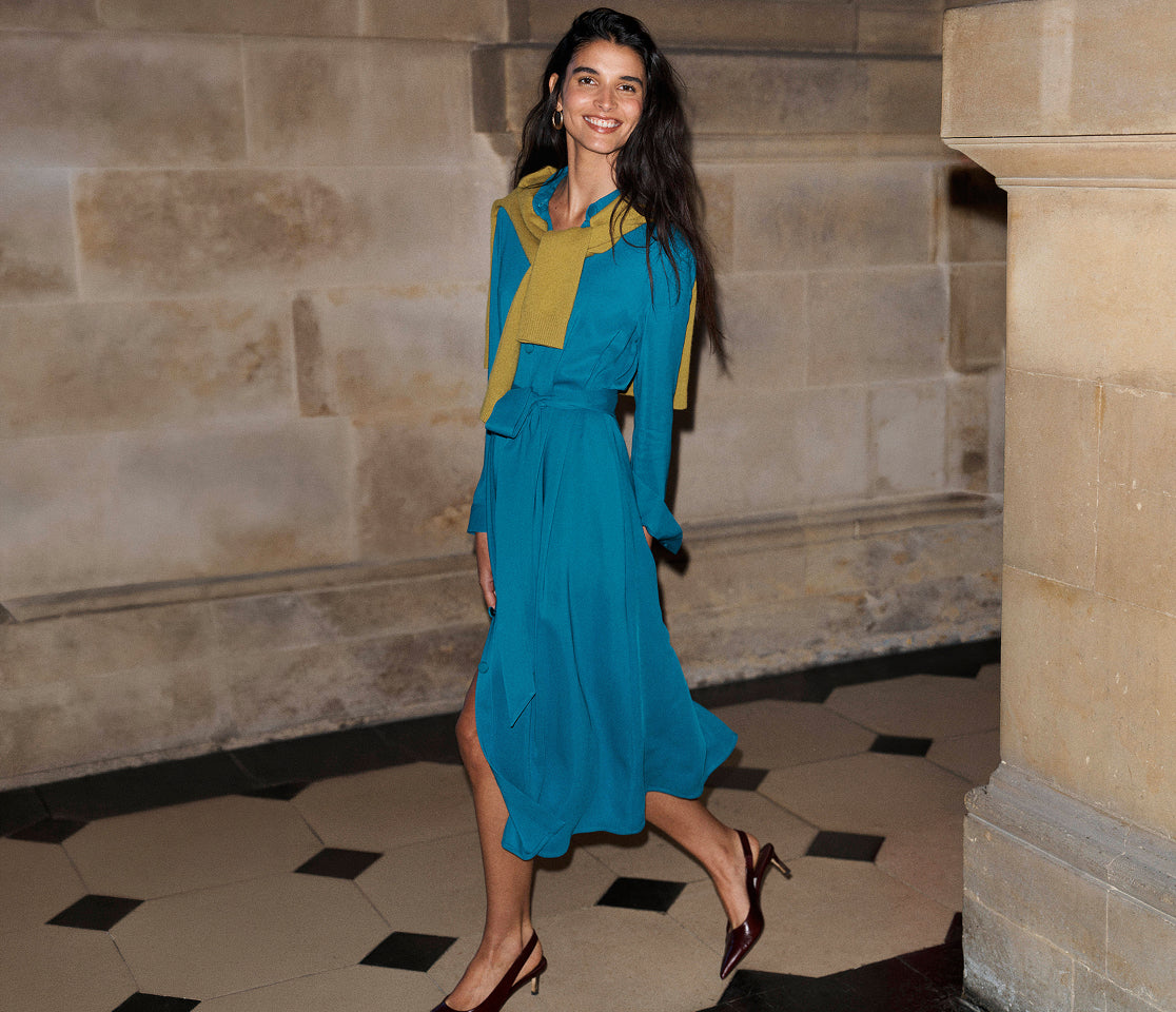A woman models a long-sleeve teal blue midi dress with a yellow accent panel on the chest and collar, styled with dark brown pointed-toe heels in an indoor stone corridor.