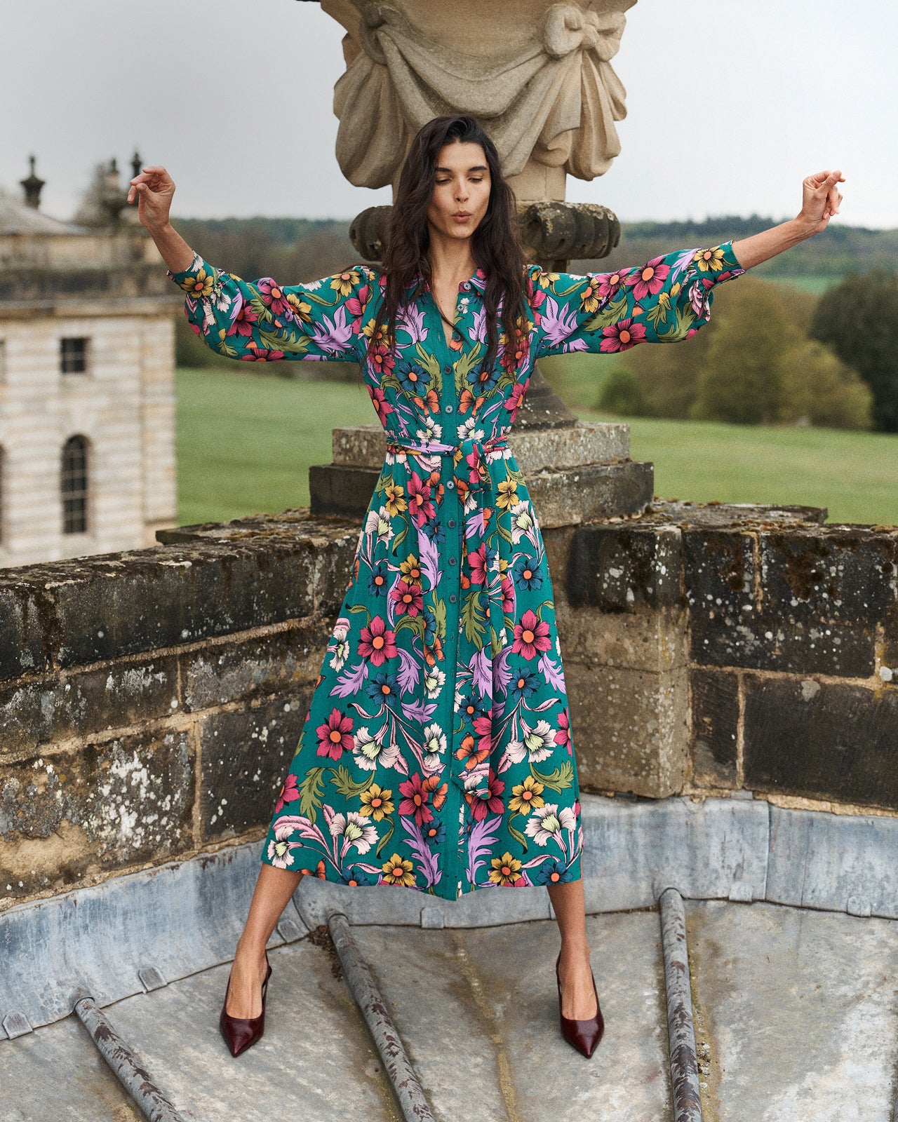 A woman stands outdoors on stone steps in front of a classical statue and green landscape, wearing a long-sleeve midi dress with a bold floral print in teal, pink, yellow, and white. She is posed with her arms outstretched, wearing dark pointed-toe heels.