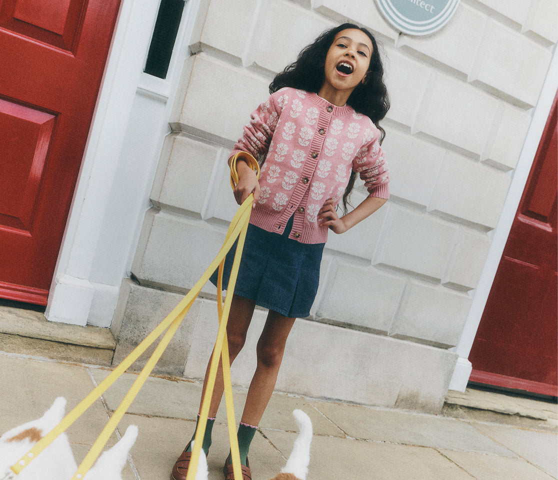 A young girl with long dark hair stands on a sidewalk in front of a white building with red doors, wearing a pink patterned cardigan, a dark blue skirt, and green socks while holding yellow dog leashes. The paws of two white dogs are visible at the bottom of the frame.