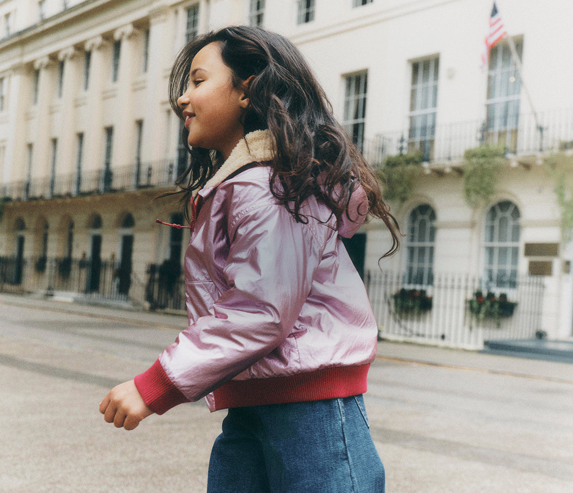 A young girl wearing a shiny light pink bomber jacket with a beige collar and red ribbed trim, walking on a city street with a classic European-style building in the background.