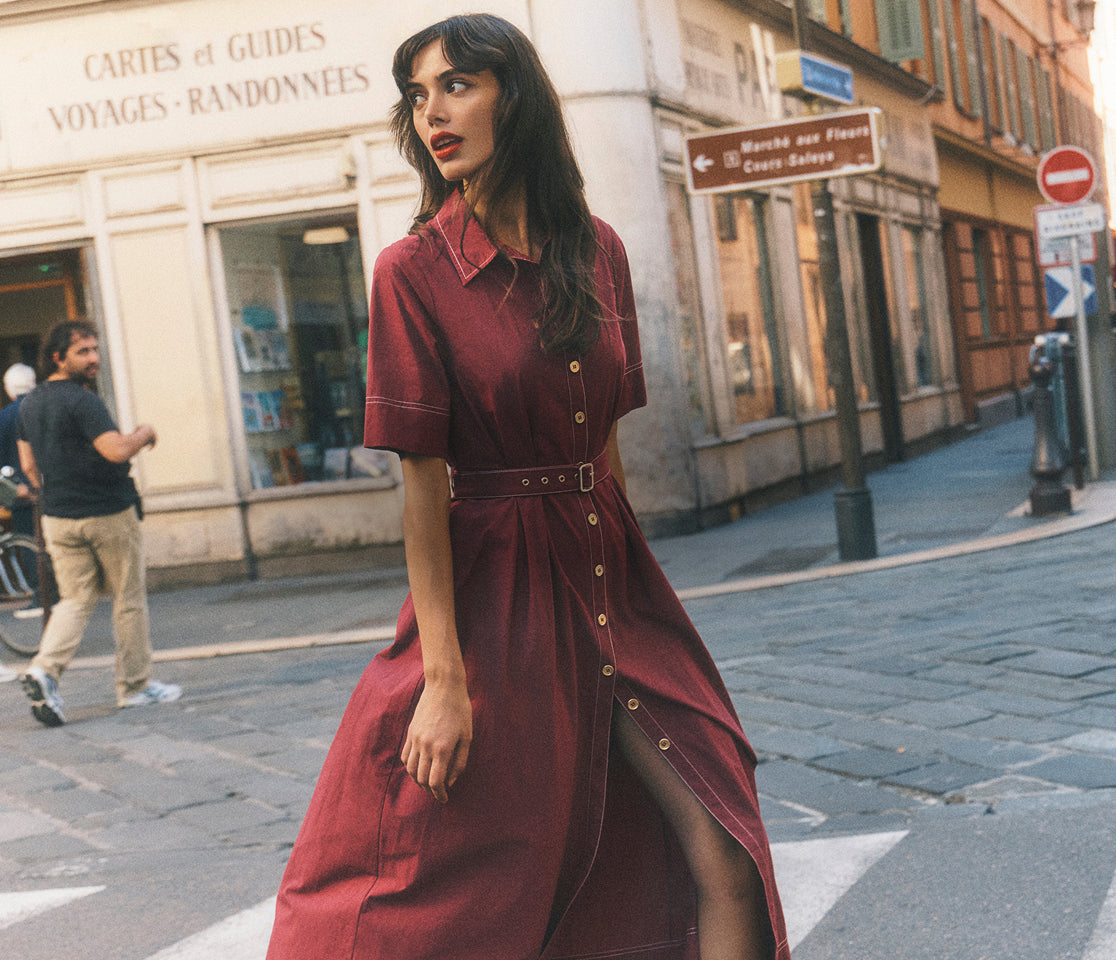 Woman in a burgundy dress walking on a street with buildings and signs in the background
