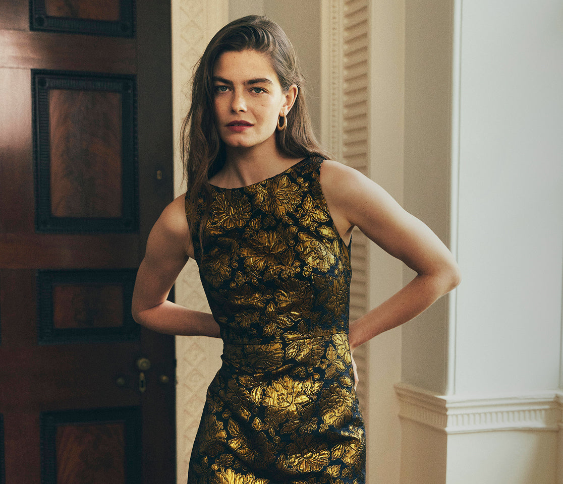 A woman modeling a sleeveless black dress with an intricate gold floral brocade pattern, standing in an elegant indoor setting with a dark wood door on the left.