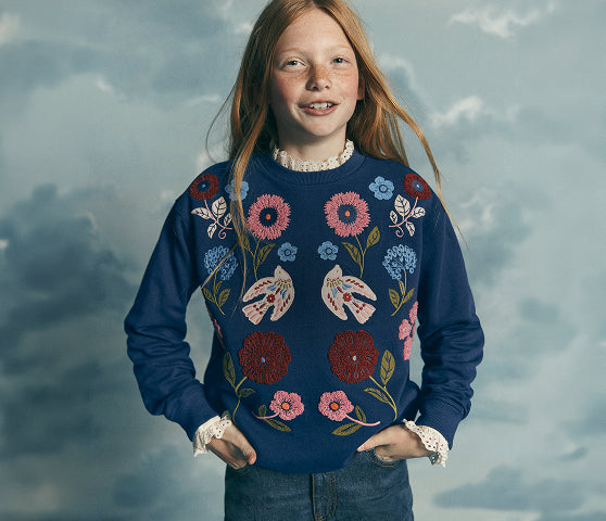 A young girl with long strawberry-blonde hair stands against a cloudy blue-gray backdrop, wearing a navy blue crewneck sweater decorated with colorful embroidered flowers and butterflies, paired with blue jeans and white lace cuffs.