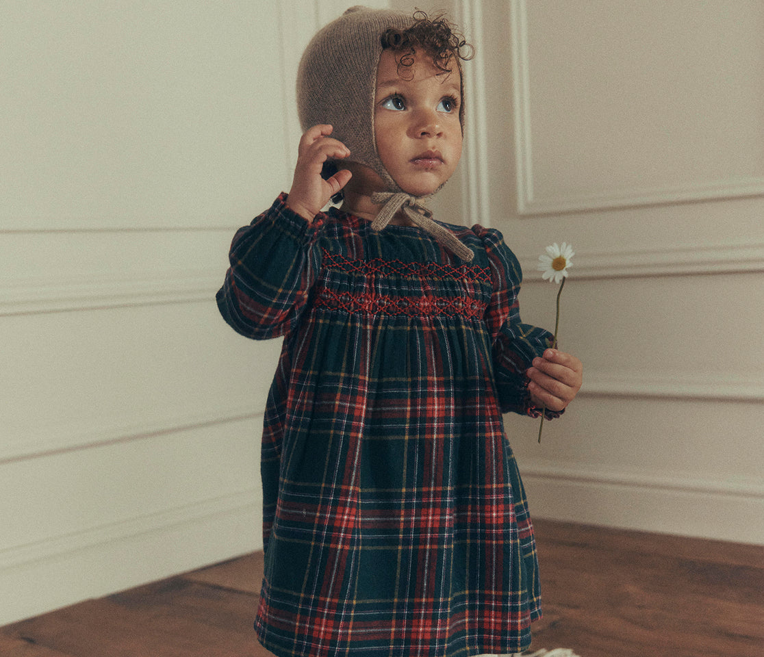 A toddler stands indoors wearing a long-sleeve plaid dress in dark green, red, and blue tones, paired with a brown knit bonnet, while holding a small white flower.