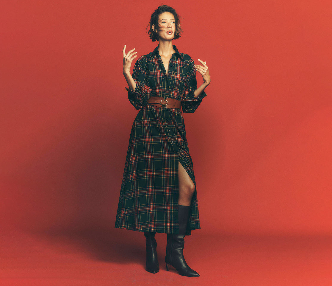 A woman modeling a long-sleeve plaid shirt dress with a brown belt and black knee-high boots, standing against a solid red studio background.