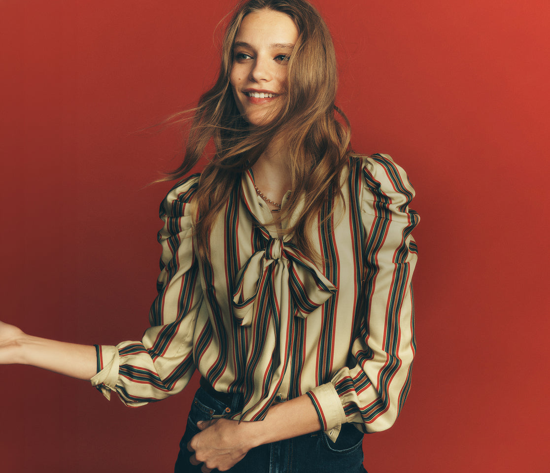 A woman with long light brown hair wearing a cream-colored blouse with vertical red, blue, and green stripes, paired with dark blue jeans, posing against a solid red background.