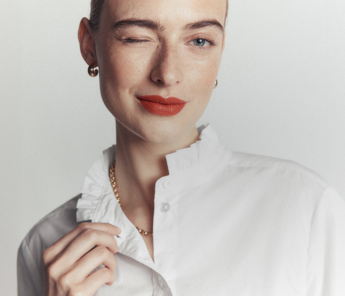 A close-up portrait of a woman wearing a white ruffled collar blouse, a delicate gold chain necklace, and a small gold stud earring, set against a plain light gray background.